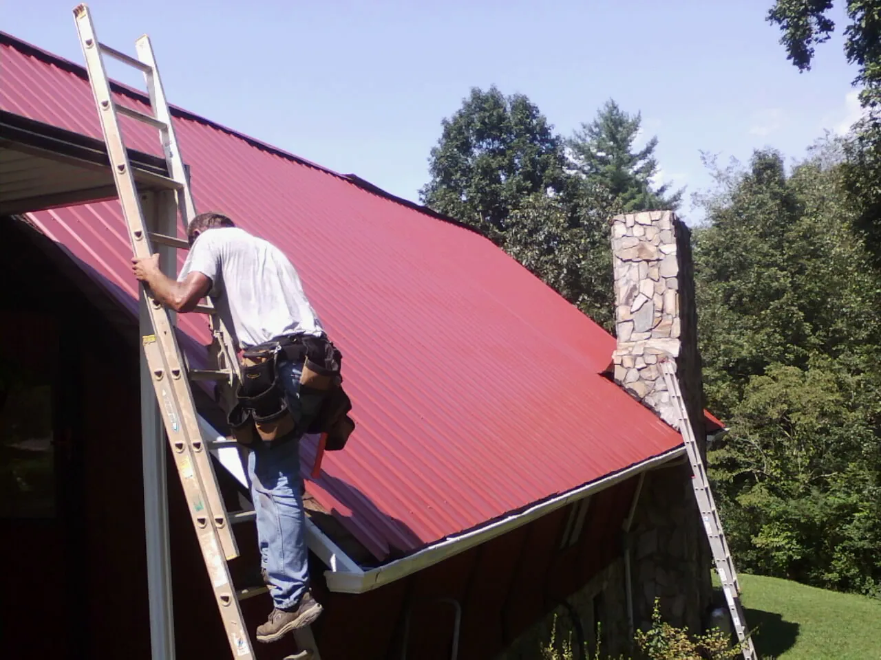 Roofer on a ladder beside a red metal roof and stone chimney — Soffit & Fascia Repair by Roofing Brothers in Rye